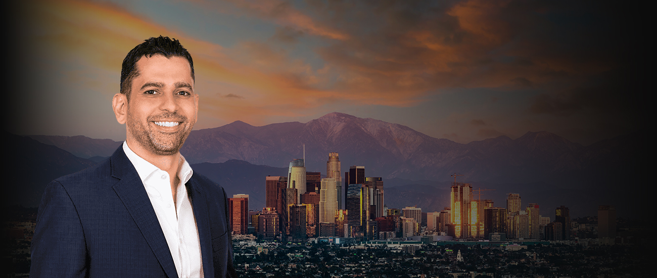 Photo of Steve Baghoomian, smiling, wearing a dark suit and a white shirt, stands in front of a blurred cityscape with skyscrapers at sunset.